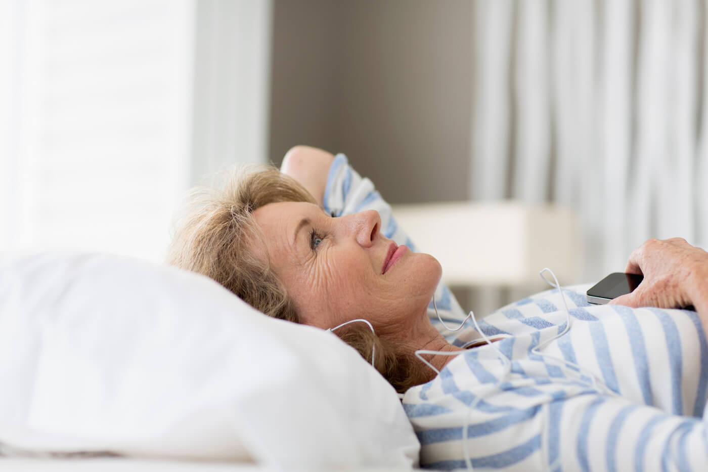 Ältere Frau liegt im Bett mit Kopfhörern und Smartphone