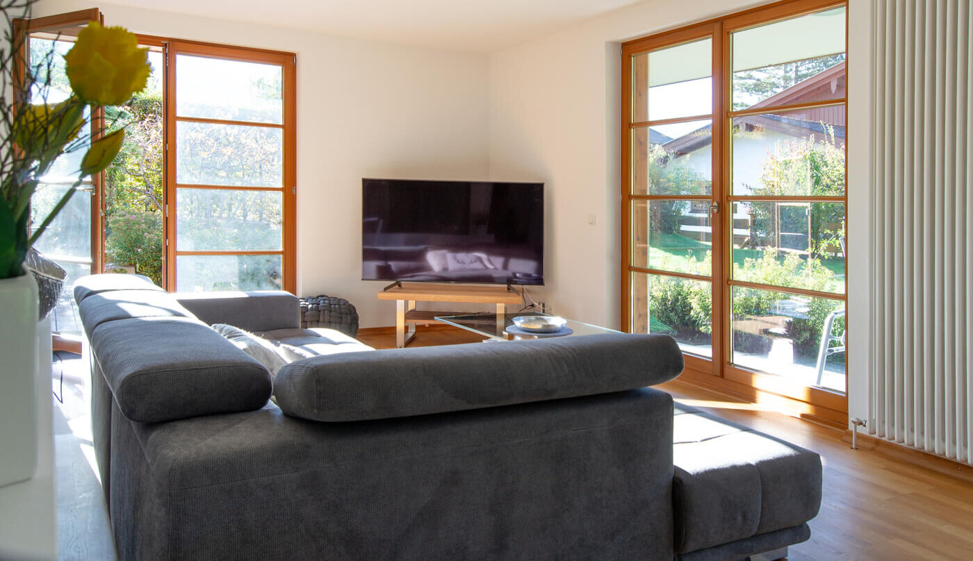 Wohnbereich der Mietwohnung 02 im Landhaus Wallbergblick in Rottach-Egern mit bodentiefen Fenstern, grauem Sofa und Blick in den Garten