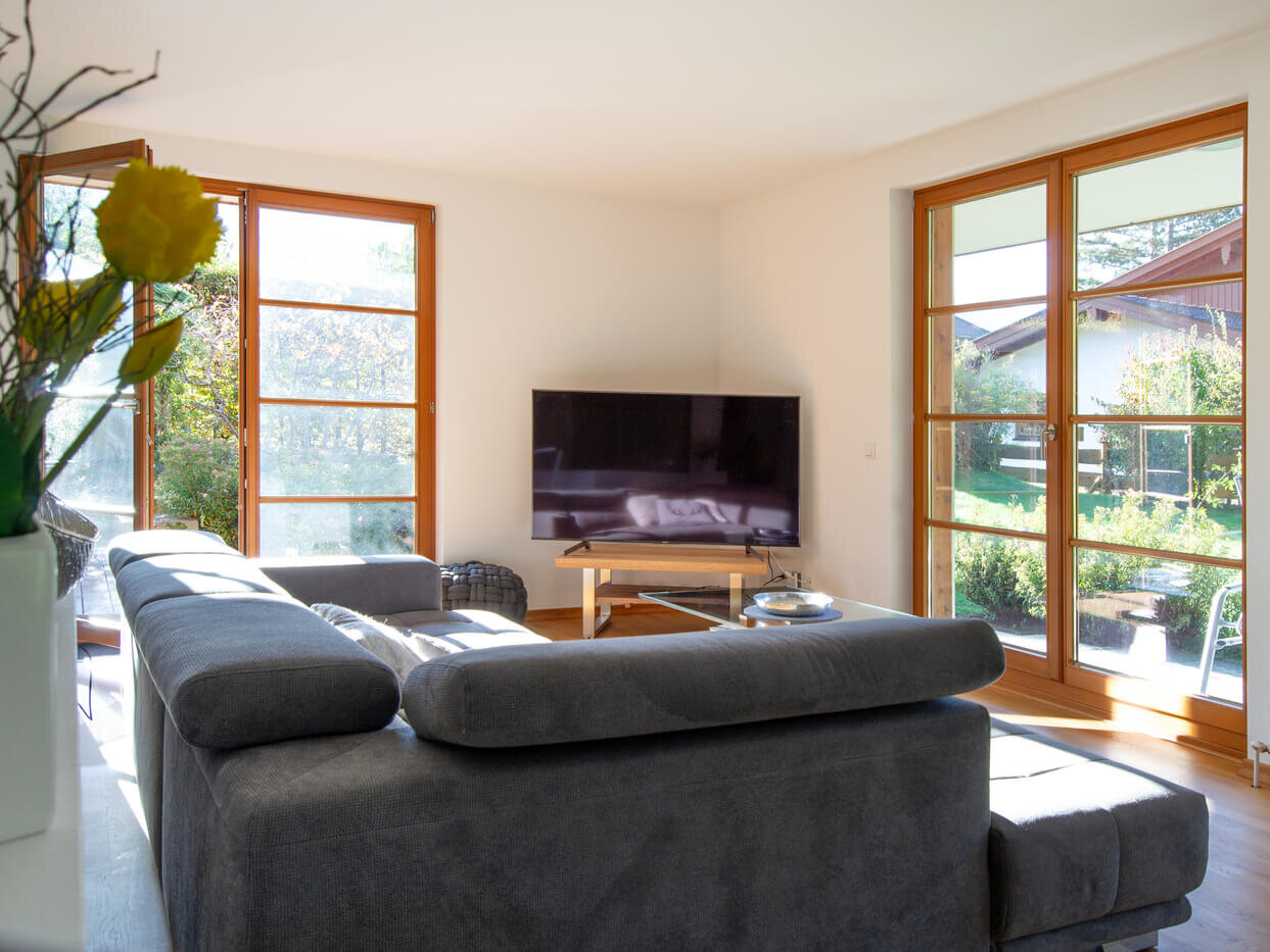 Wohnbereich der Mietwohnung 02 im Landhaus Wallbergblick in Rottach-Egern mit bodentiefen Fenstern, grauem Sofa und Blick in den Garten